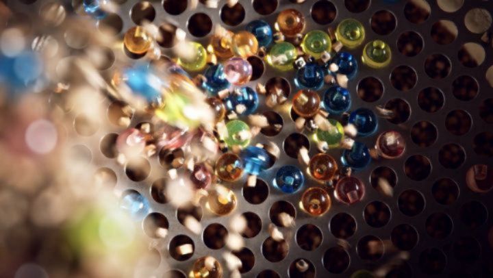 Close-up of colorful beads dropping through holes of a metal strainer, showing motion blur and selective focus.