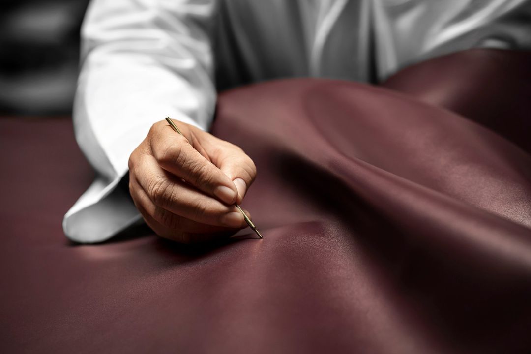 Close-up shot of a person in a white lab coat using a fine-point tool on dark red leather, highlighting craftsmanship and precision.