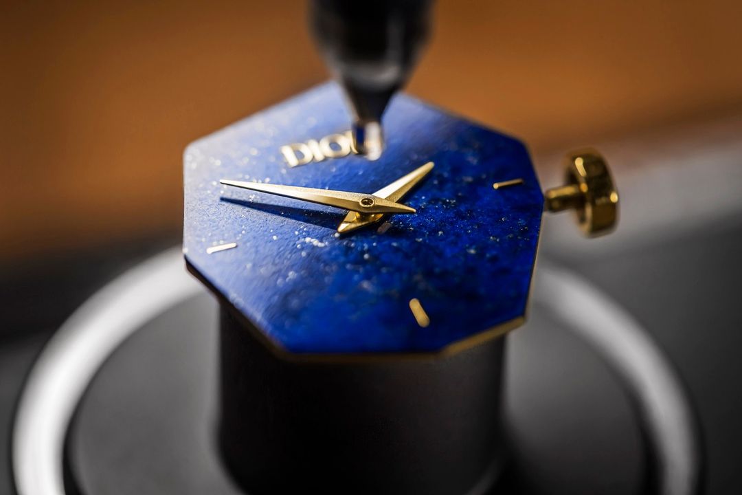 Close-up of a Dior watch featuring gold hands and a blue face with speckled details, set against a blurred background.