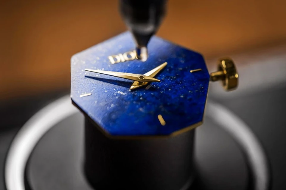 Close-up of a Dior watch featuring gold hands and a blue face with speckled details, set against a blurred background.