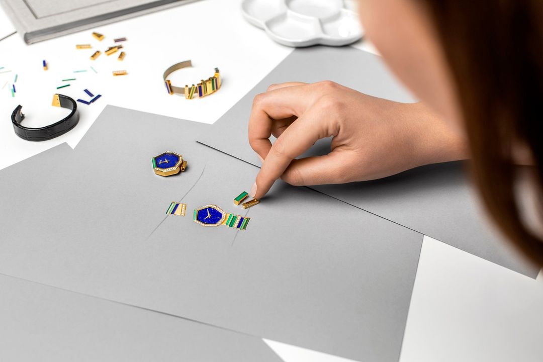 Woman designs custom watch with precious stones and gold elements on a grey surface in an indoor setting with soft, diffused lighting.