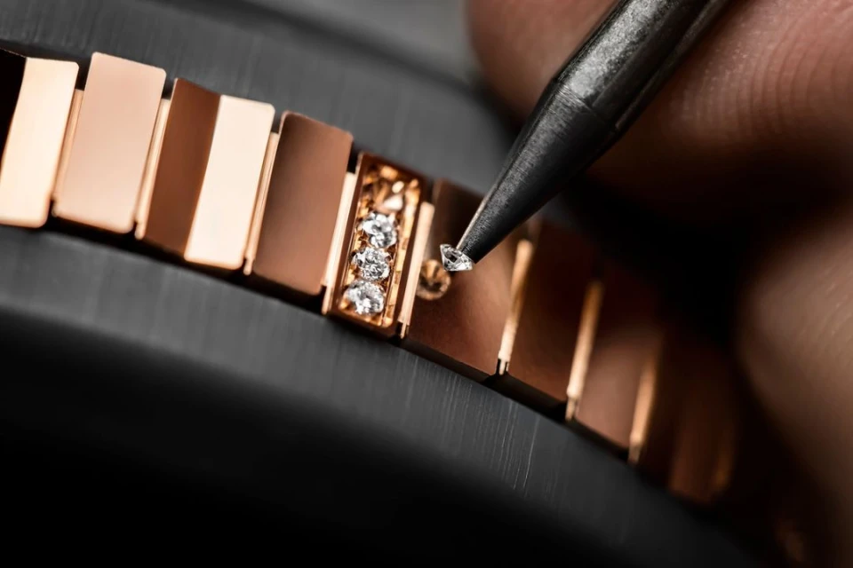 Close-up shot of a jeweller setting a diamond on a rose-gold watch bracelet with a small tool, against a dark grey background.
