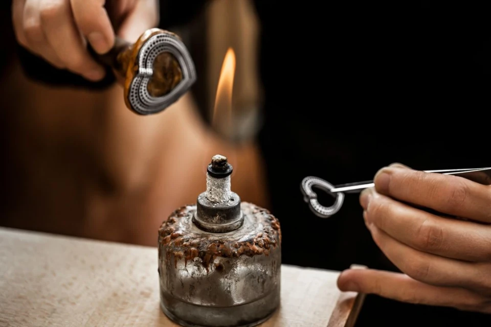 Close-up of a jewellery maker using a small torch to heat metal for design, with hands and tools visible, on a workbench.