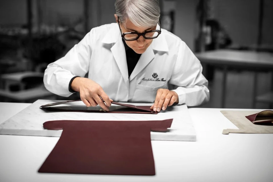An adult woman with short grey hair wearing a white lab coat is working on a burgundy leather product in an indoor environment.