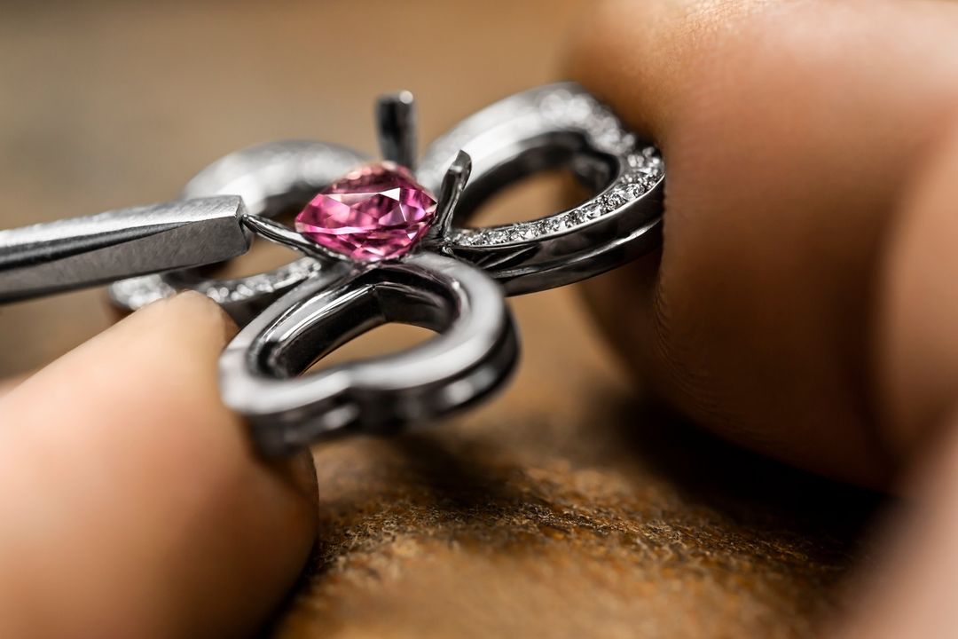Close-up of a pink gemstone being set in a silver ring with small diamonds using a pair of metal tweezers on a wooden surface.