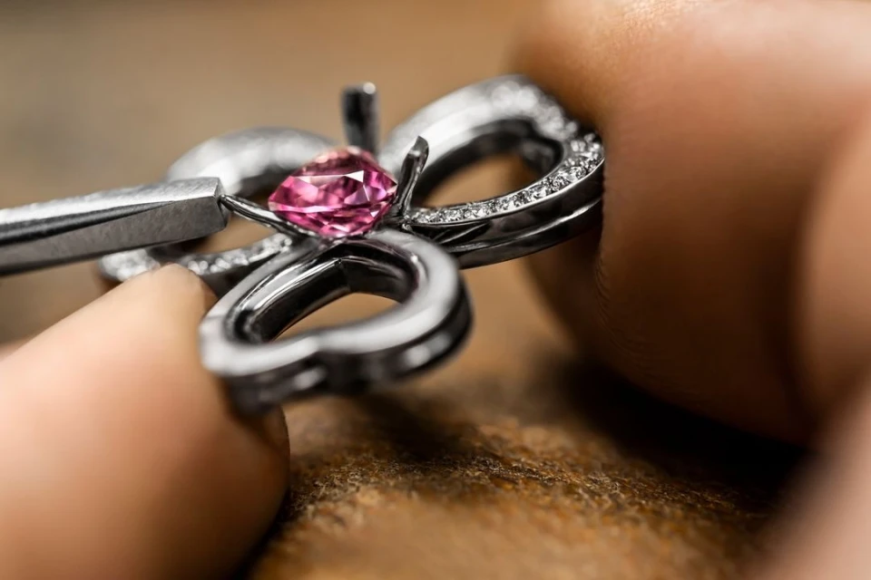 Close-up of a pink gemstone being set in a silver ring with small diamonds using a pair of metal tweezers on a wooden surface.
