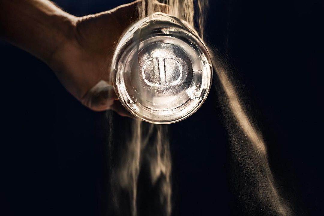 Close-up of a hand holding a clear glass bottle with sand pouring out of it against a dark background in a studio setting.