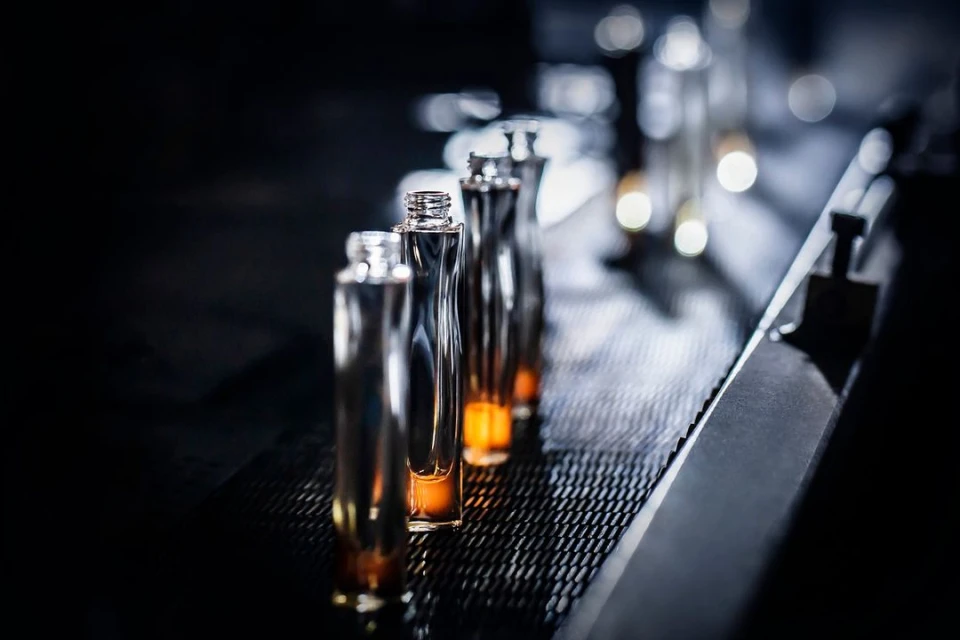 Several clear glass bottles move along a dark conveyor belt, with a blurred background, suggesting an industrial or manufacturing setting.