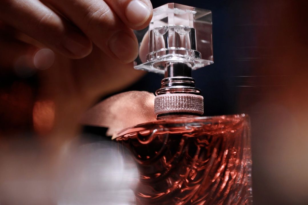 Close-up shot of a person's hand placing a clear, square cap on top of an ornately patterned rose-colored glass bottle.