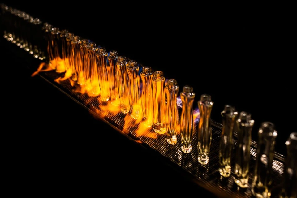 Rows of transparent glass vials are lit by flames on a reflective surface against a dark backdrop, showcasing their uniformity.