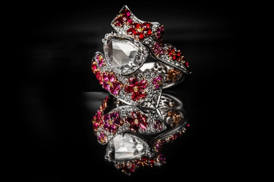 Close-up shot of an ornate silver ring adorned with a clear stone and red and white gems, displayed on a reflective black surface.