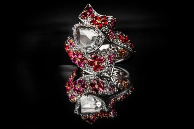 Close-up shot of an ornate silver ring adorned with a clear stone and red and white gems, displayed on a reflective black surface.