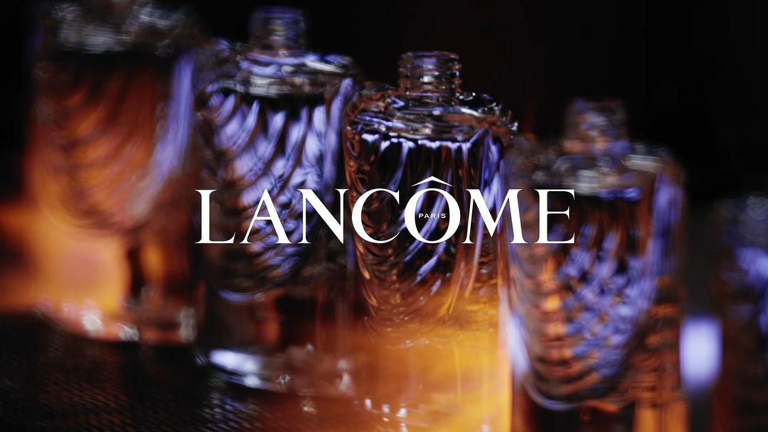 Close-up of Lancôme perfume bottles with abstract design, lit with amber and purple hues against a dark background, evoking luxury.