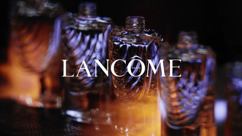 Close-up of Lancôme perfume bottles with abstract design, lit with amber and purple hues against a dark background, evoking luxury.