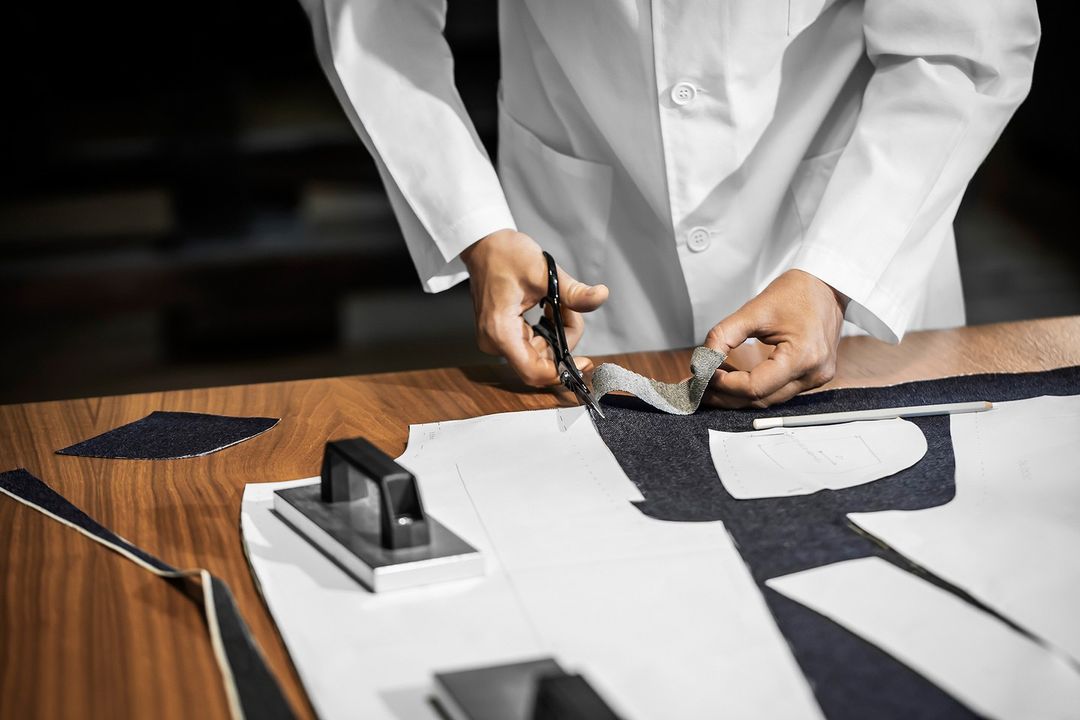 A tailor cuts grey material with scissors on a wooden table, surrounded by white paper patterns and tools, in a crafting environment.