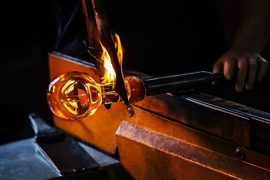 A glassblower shapes molten glass with fire, using a metal tool in a dark workshop setting with vibrant orange and yellow hues.