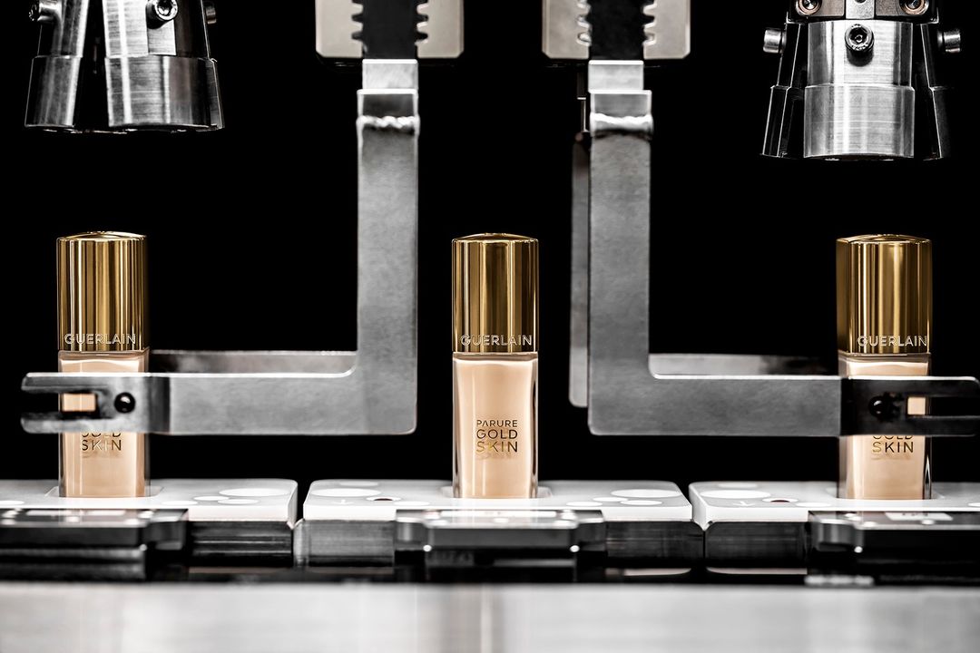 Close-up product shot of Guerlain Parure Gold Skin foundation bottles in tan and gold, centered between metallic machinery in studio setting.
