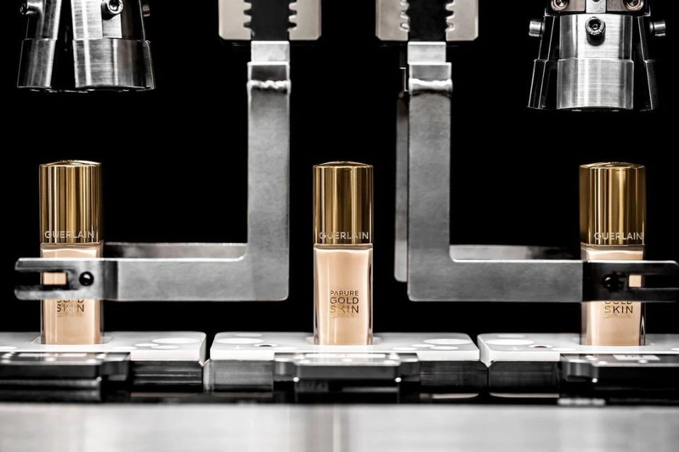 Close-up product shot of Guerlain Parure Gold Skin foundation bottles in tan and gold, centered between metallic machinery in studio setting.