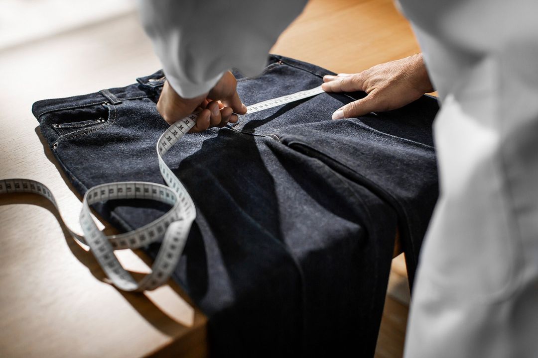 Denim jeans are being measured with a white measuring tape on a wooden table, with a person in a white coat standing behind.