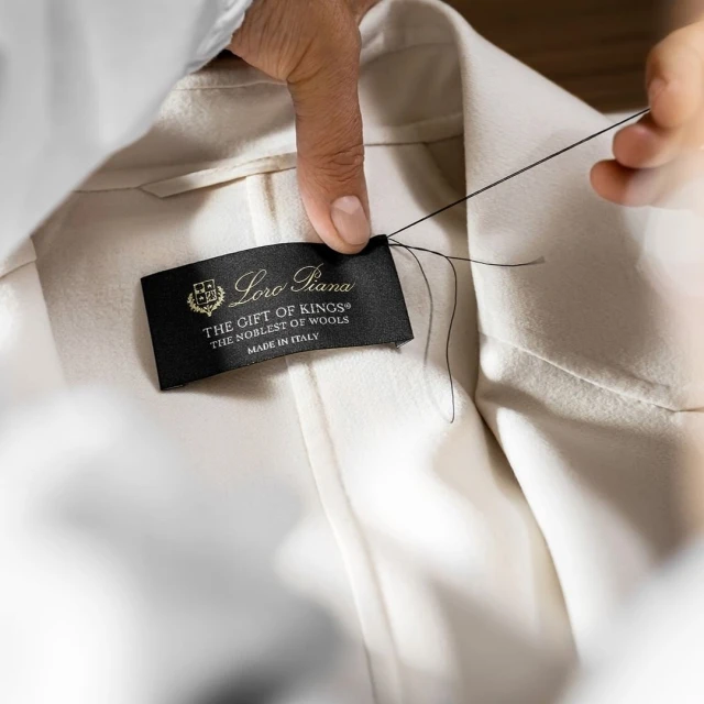 Close-up of a tailor's hand sewing a 'Loro Piana' brand label onto a garment, showcasing craftsmanship and high-end fashion detailing.