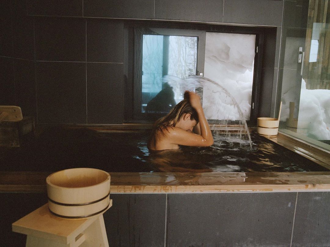 Woman relaxes in a dark tiled indoor bath with running water near a window with snowy outdoor scenery, creating a serene and tranquil scene.