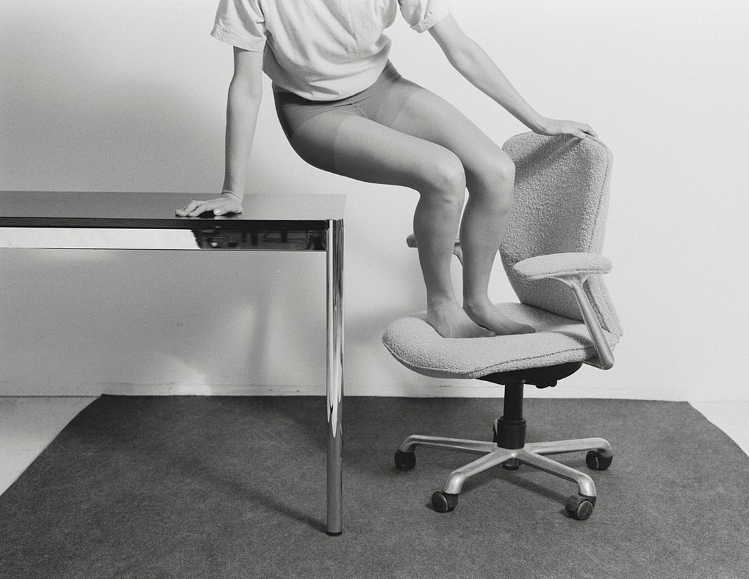 Monochromatic studio shot of an adult standing barefoot on an office chair next to a metal table, minimalist interior and design.