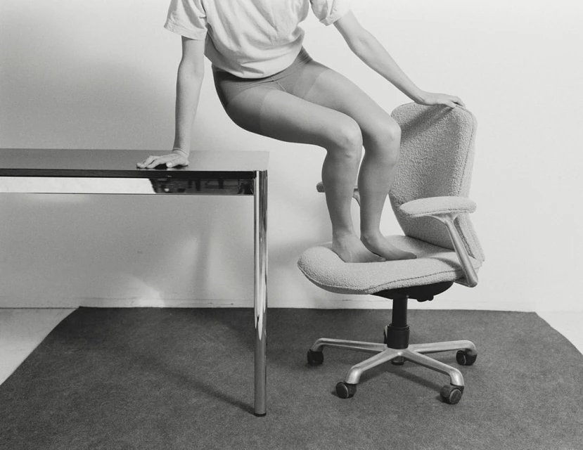Monochromatic studio shot of an adult standing barefoot on an office chair next to a metal table, minimalist interior and design.