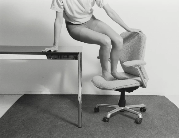 Monochromatic studio shot of an adult standing barefoot on an office chair next to a metal table, minimalist interior and design.