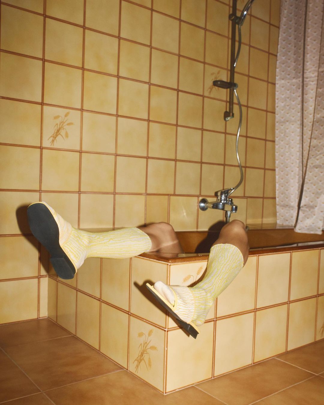 A person's lower body is submerged in a tiled bathtub, wearing yellow socks and dress shoes, in a tiled bathroom with a shower.
