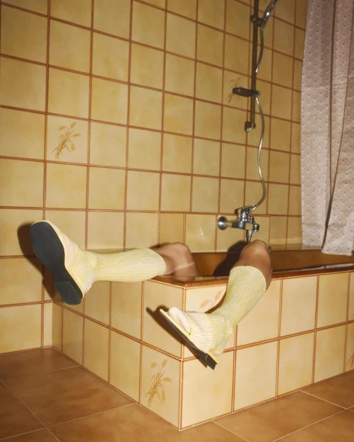 A person's lower body is submerged in a tiled bathtub, wearing yellow socks and dress shoes, in a tiled bathroom with a shower.
