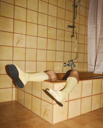 A person's lower body is submerged in a tiled bathtub, wearing yellow socks and dress shoes, in a tiled bathroom with a shower.