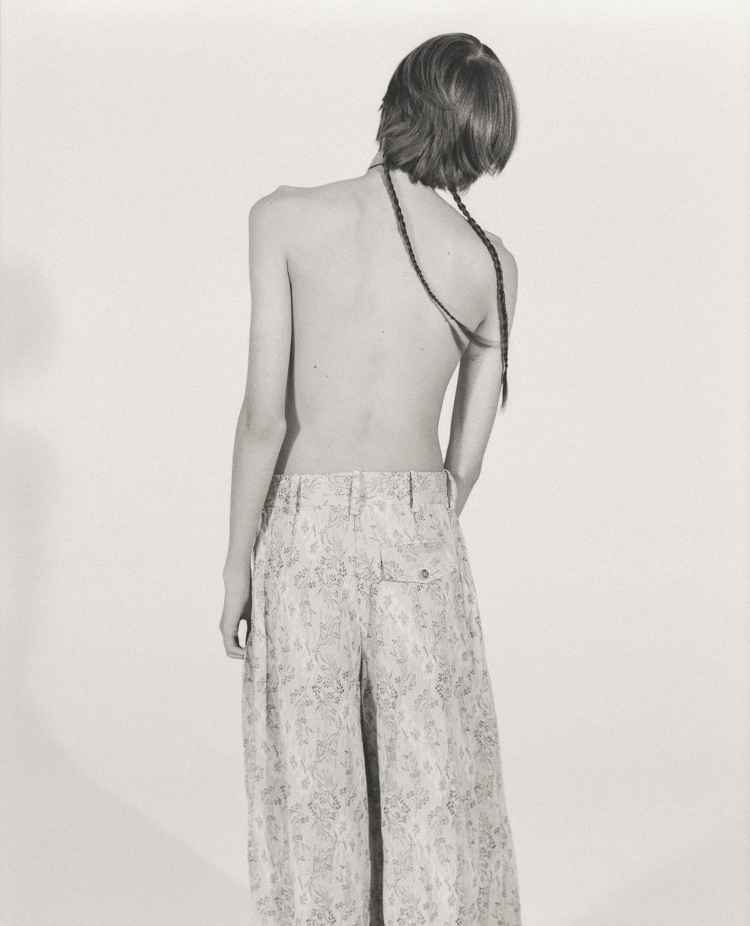 Back view of a slender person with braided hair, wearing patterned palazzo pants, standing in a monochrome indoor studio.