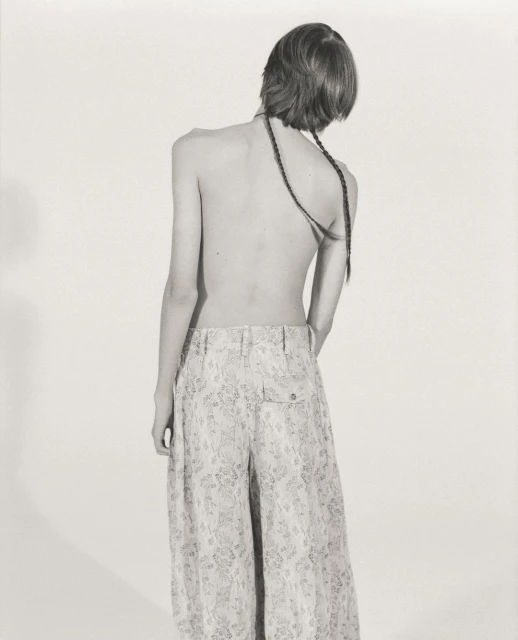 Back view of a slender person with braided hair, wearing patterned palazzo pants, standing in a monochrome indoor studio.