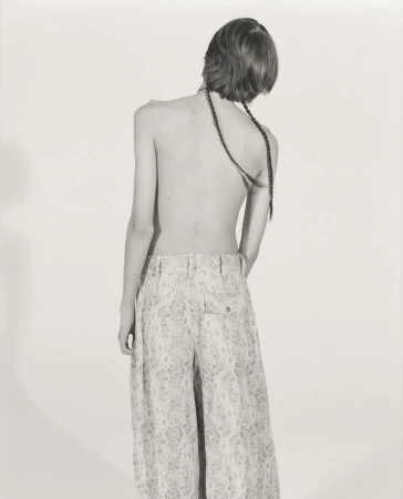Back view of a slender person with braided hair, wearing patterned palazzo pants, standing in a monochrome indoor studio.