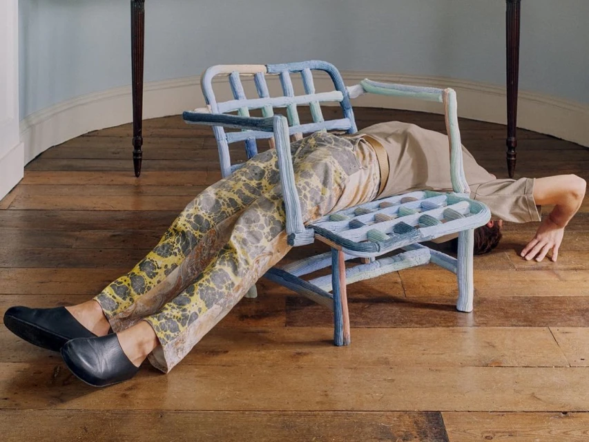 Person lying on a blue woven chair in a room with hardwood floors and a light blue wall, creating a surreal and artistic scene.