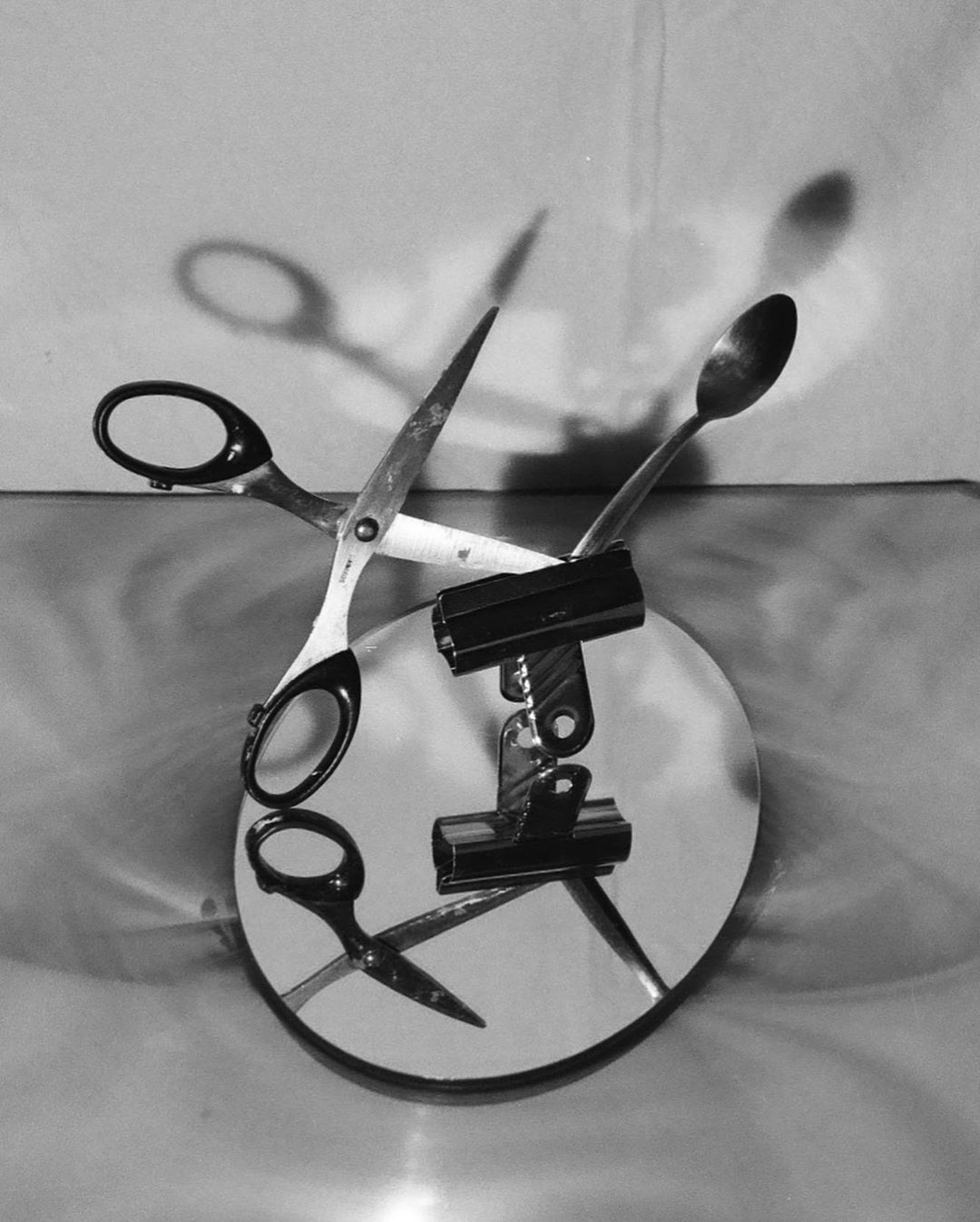 Still life of scissors, binder clips, and a spoon on a round mirror with a light-colored background and shadows. Black and white image.