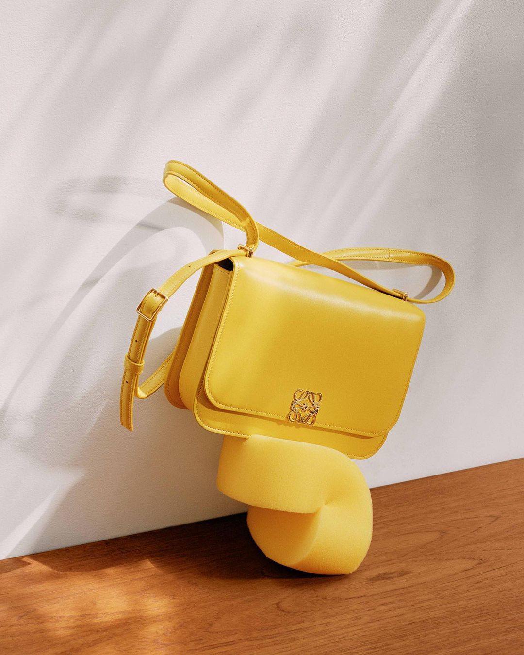 Elevated view of a yellow Loewe handbag on a yellow plinth against a textured white wall and wood floor, showcasing a studio product shot.