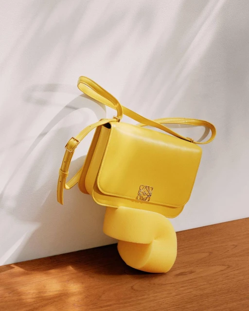 Elevated view of a yellow Loewe handbag on a yellow plinth against a textured white wall and wood floor, showcasing a studio product shot.
