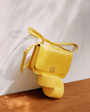 Elevated view of a yellow Loewe handbag on a yellow plinth against a textured white wall and wood floor, showcasing a studio product shot.