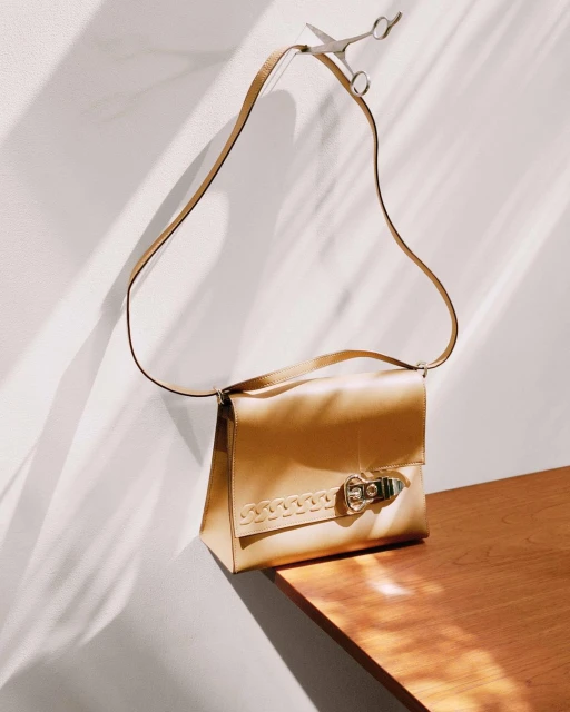 Gold satchel handbag with shoulder strap is suspended from a pair of scissors against a neutral wall with light and shadow play.