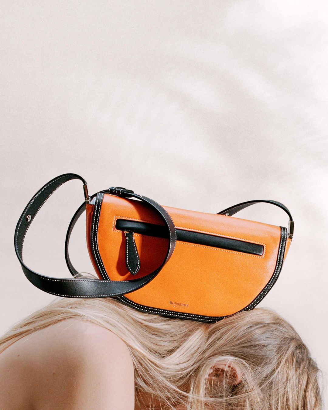 Orange leather shoulder bag with black trim and strap, resting on a person's head with blond hair against a light neutral backdrop.