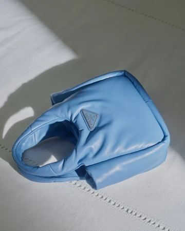 Close-up of a padded blue leather handbag resting on a white fabric surface with soft natural light.
