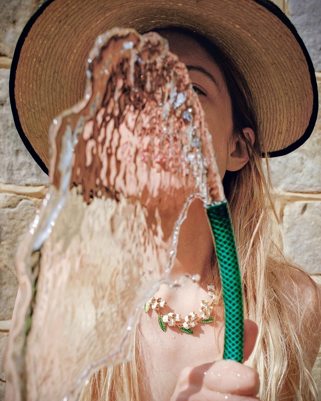 A woman in a straw hat holds a green garden hose, spraying water towards the camera. She is wearing a floral necklace.