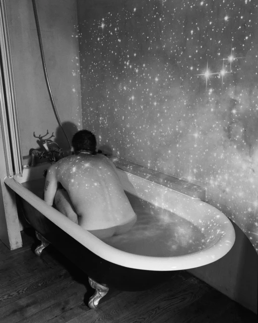 Monochrome image of a shirtless adult in a clawfoot bathtub, with a starry sky projected on the wall behind in an indoor setting.