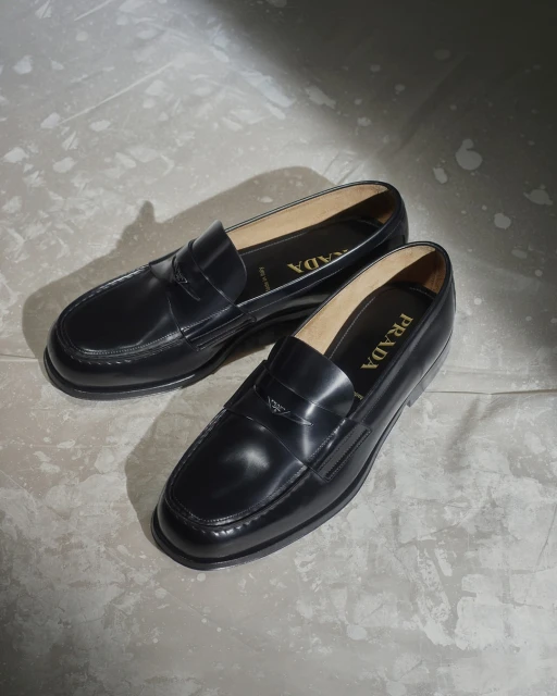 Close-up of a pair of black leather Prada loafers on a textured grey surface, bathed in soft natural light.