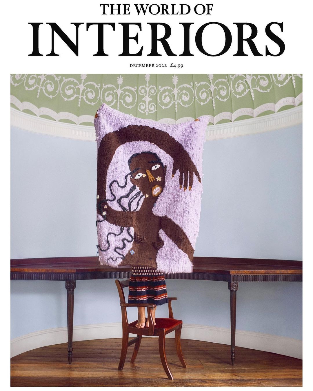 Magazine cover featuring a dark-skinned person in a fringed skirt standing on a chair behind a purple tapestry, with a table nearby.