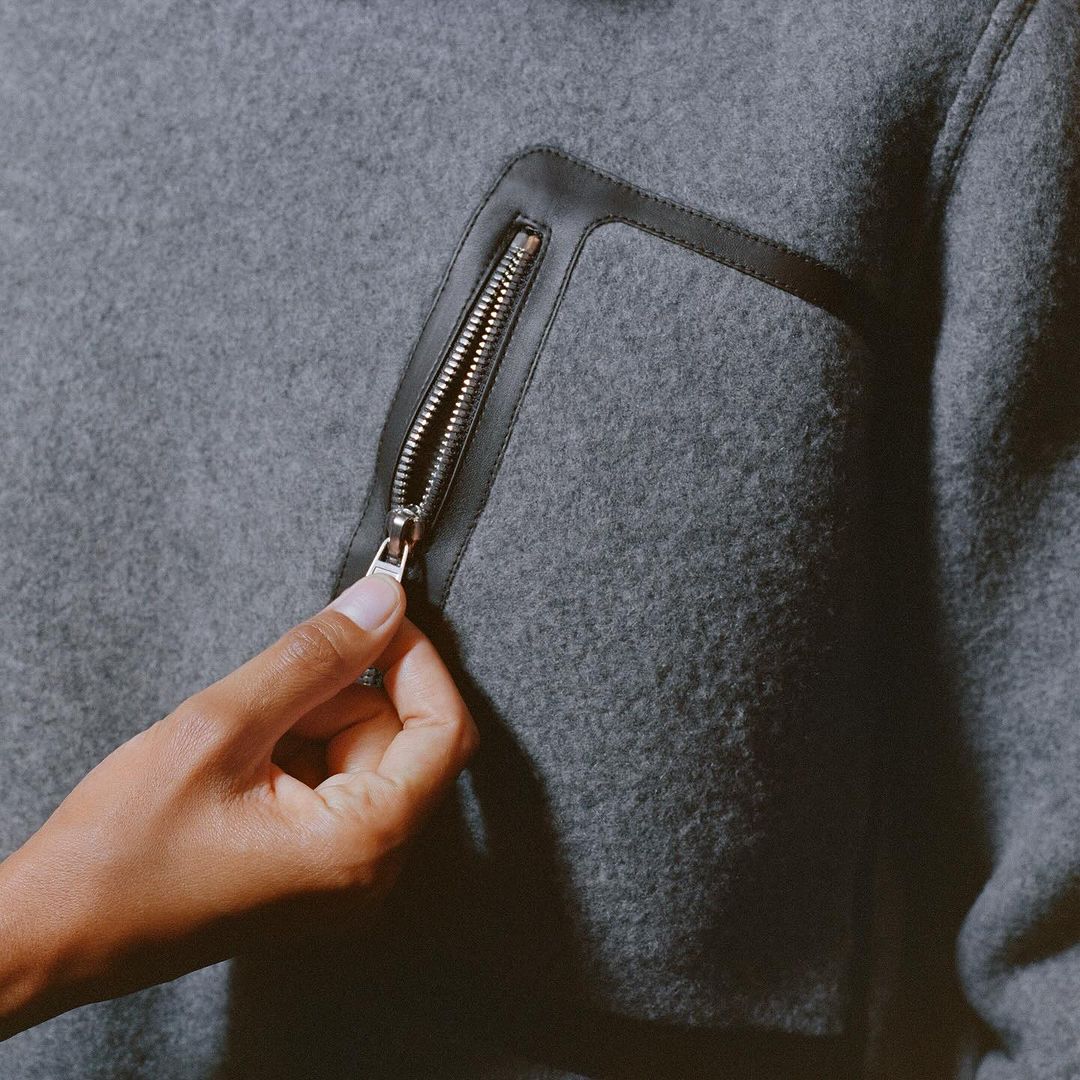 A close-up of a grey wool jacket with a black leather pocket trim and a hand partially opening the silver zipper.
