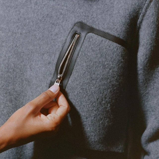 A close-up of a grey wool jacket with a black leather pocket trim and a hand partially opening the silver zipper.