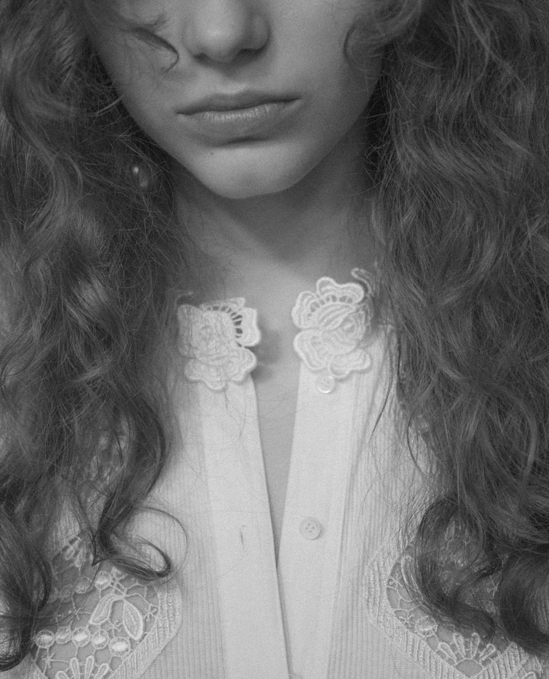 Close-up black and white shot of a young woman with wavy hair wearing a white shirt with lace detailing and decorative collar clasps.
