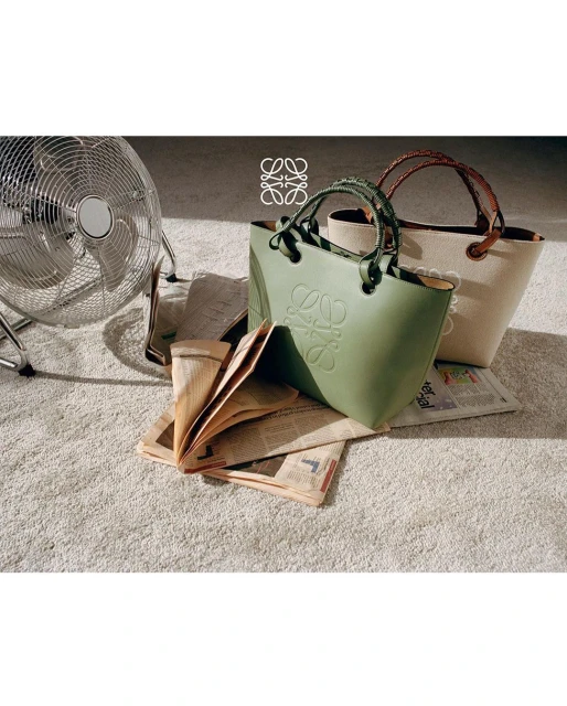 Loewe Puzzle Fold tote bags in green and beige displayed on a carpet with a floor fan and newspapers in the background.